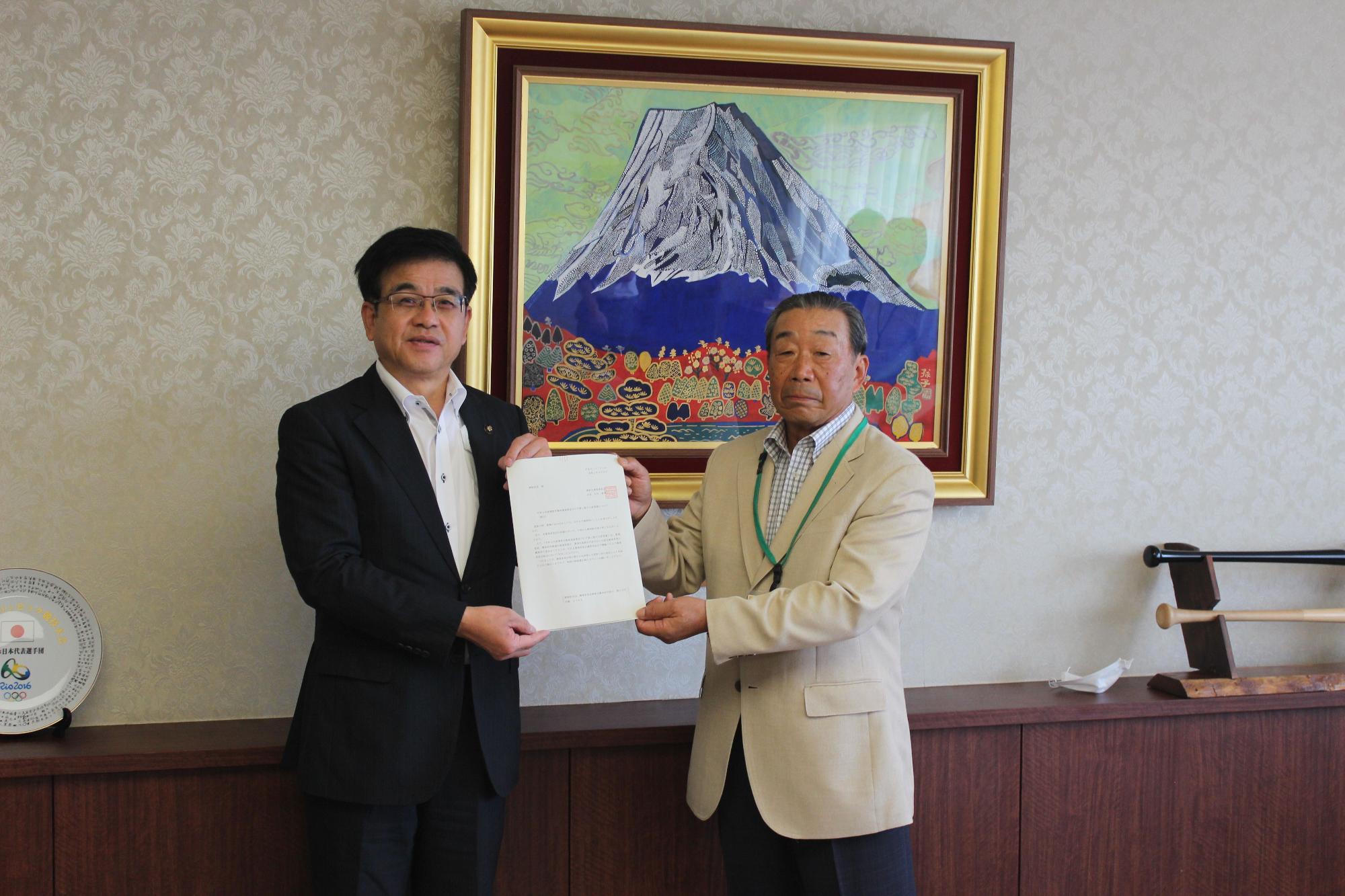 富士山の絵画が飾られた室内で、スーツ姿の市長と男性が、中央で要望書を並んで持っている写真