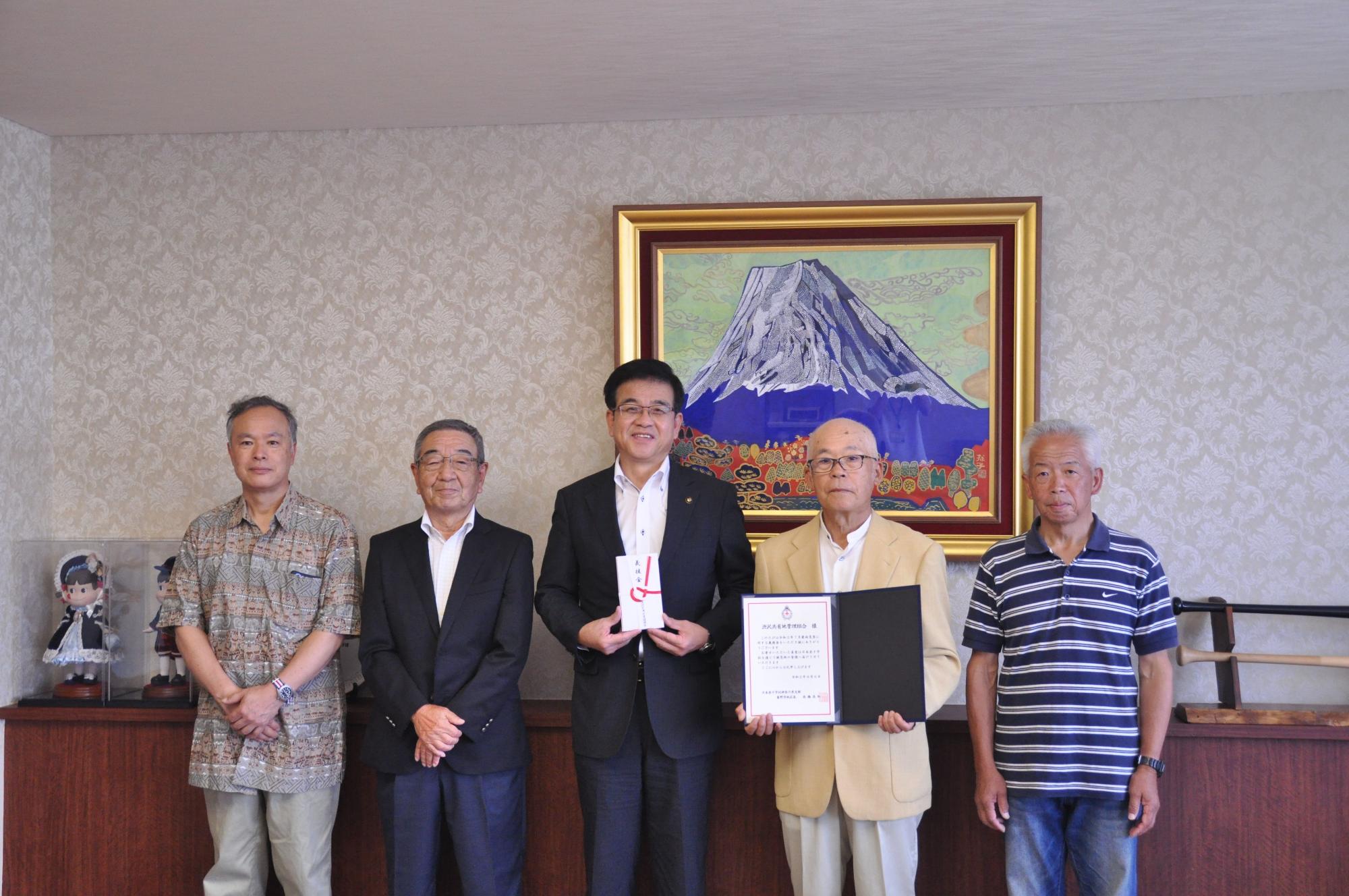 富士山の絵画が飾られた部屋で、市長を含む5人の男性が立ち、中央の市長は紅白の熨斗紙の目録、その隣の男性は感謝状のような書類を持ち並んでいる写真