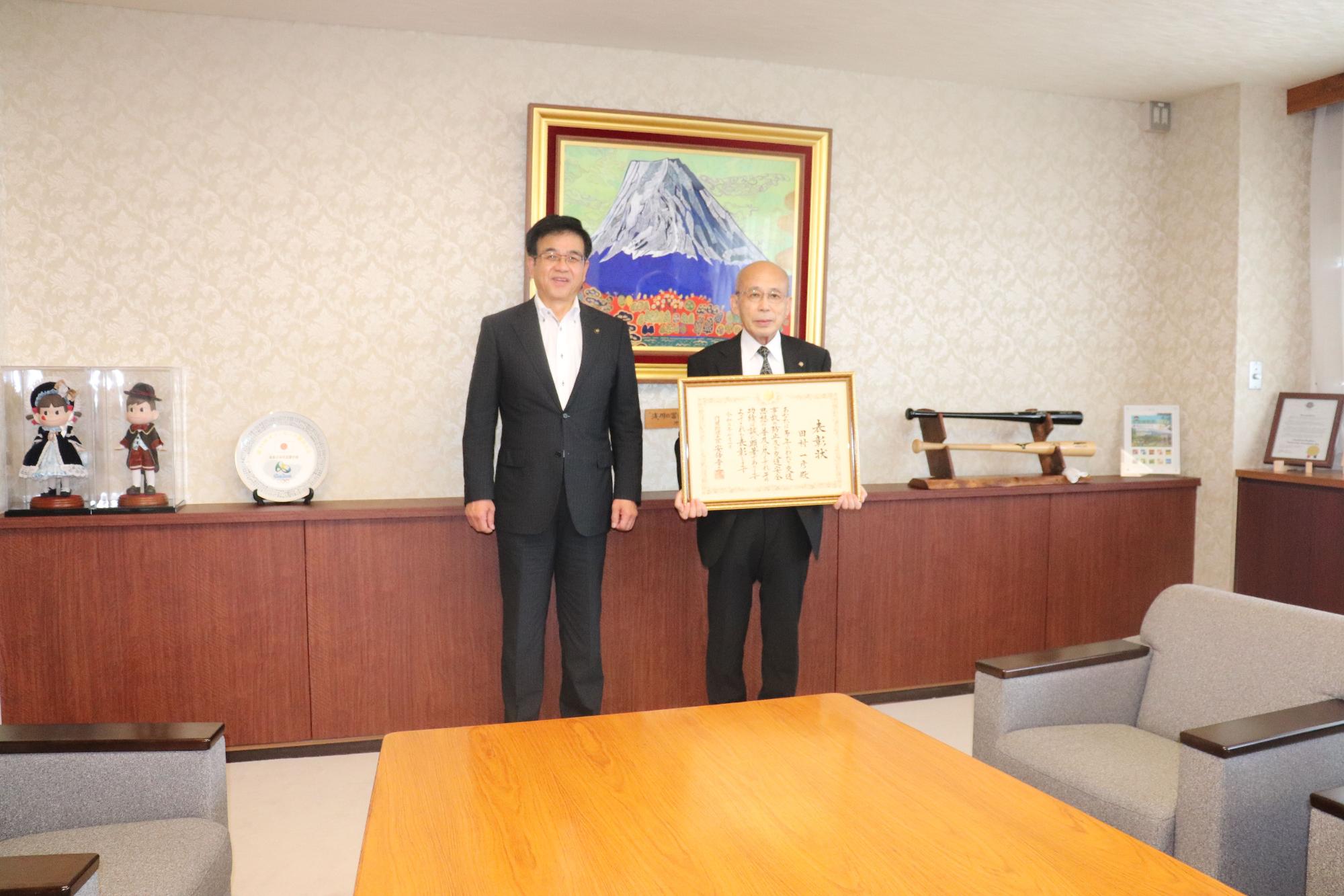 富士山の絵画が飾られた部屋で、市長と額縁入りの賞状を持った男性が立っている写真