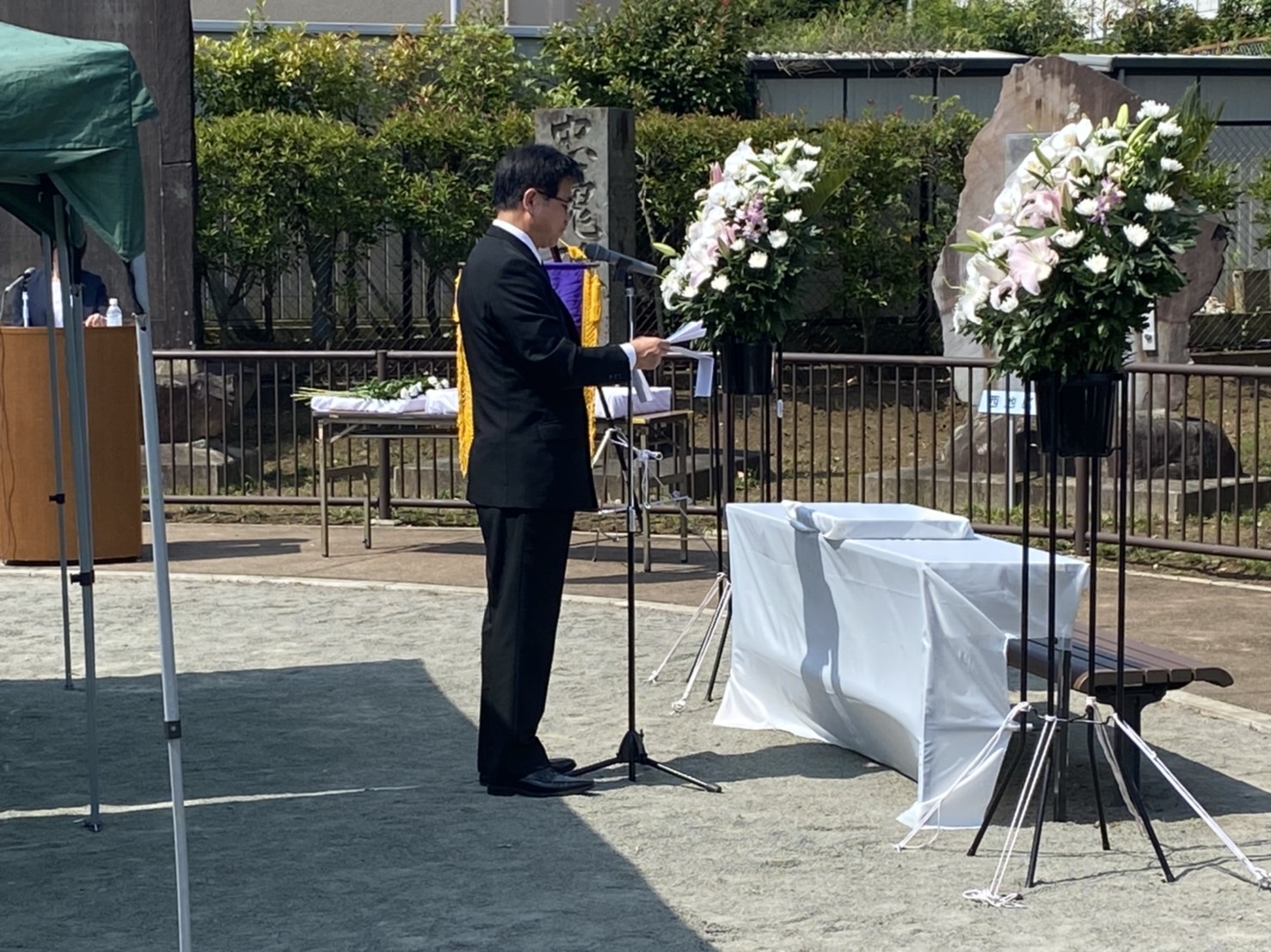 晴れた屋外で、白とピンクの生花が飾られた祭壇の前で、スーツ姿の市長がマイクスタンドの前で式辞を読み上げている追悼式典の写真