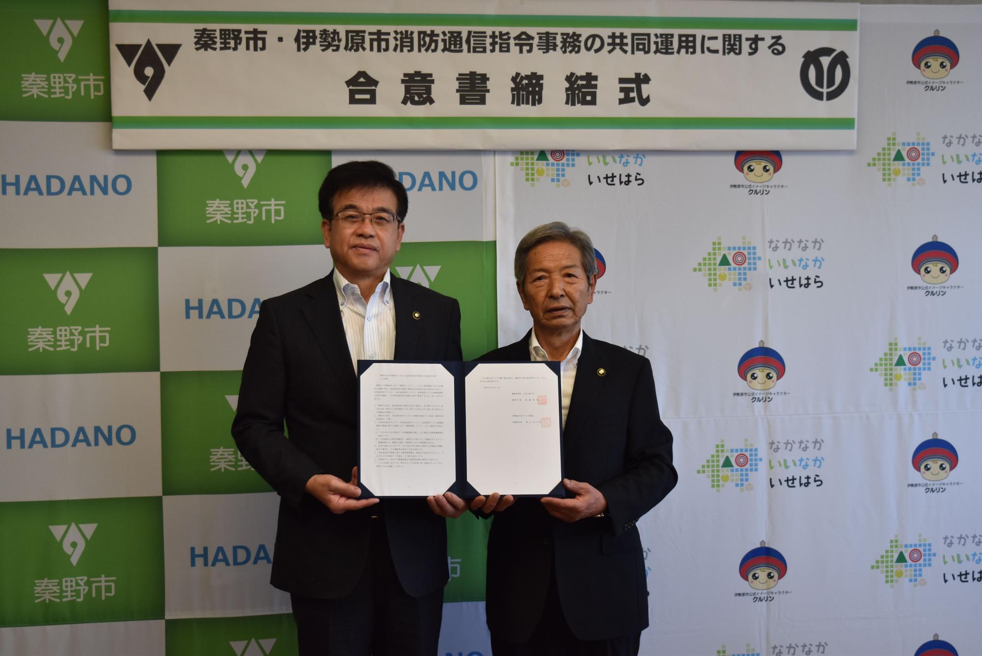 秦野市と伊勢原市の市長が、「秦野市・伊勢原市消防通信指令事務の共同運用に関する合意書締結式」の看板の前で、開かれた合意書を挟んで立っている記念写真