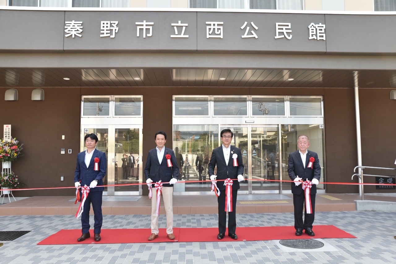濃い茶色の建物正面の「秦野市立西公民館」の文字の下で、紅白のテープにハサミを入れる市長と3人の男性を、赤いカーペット越しに写したテープカットの写真