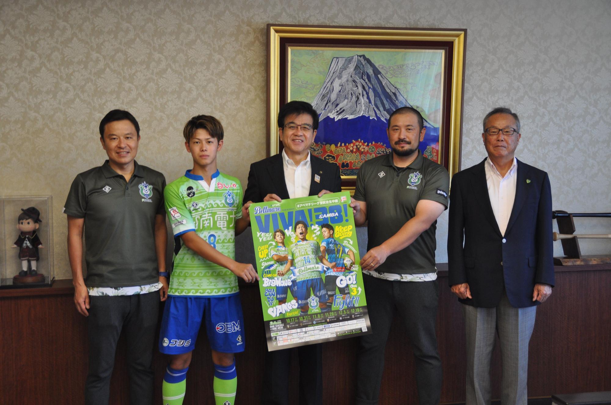 富士山の絵画が飾られた室内で、ユニフォーム姿のサッカー選手と市長と関係者を含む5人の男性が、中央でポスターを広げて記念撮影をしている写真