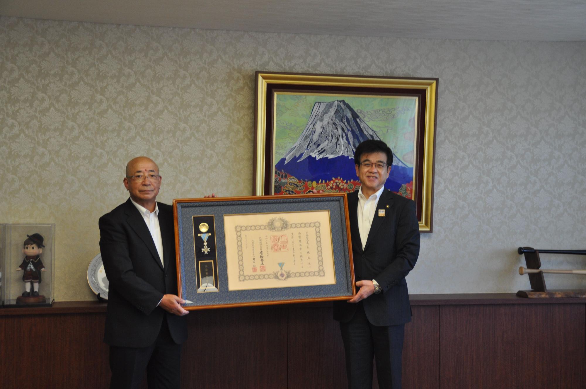 富士山の絵画が飾られた室内で、スーツ姿の市長と男性が、メダルと写真が添えられた大きな額縁入りの賞状を並んで持っている写真