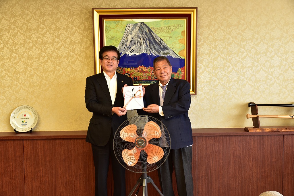 富士山の絵画が飾られた室内で、スーツ姿の市長と男性が、中央で白い熨斗袋の目録を扇風機越しに持って立っている写真