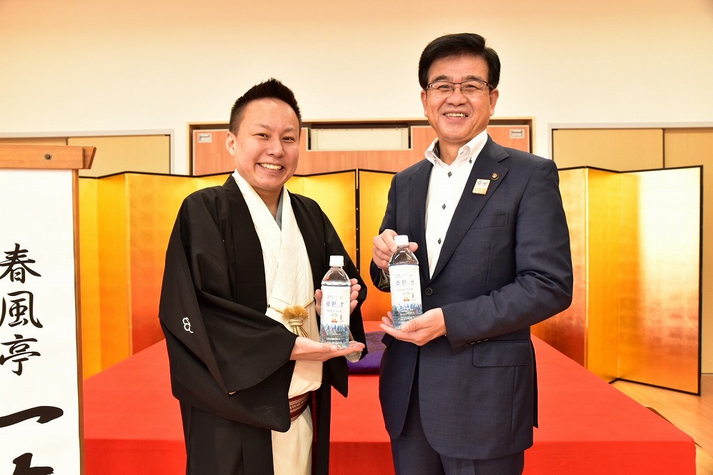 赤い毛氈が敷かれた高座の前で、黒い着物姿の男性とスーツ姿の市長が、それぞれミネラルウォーターのペットボトルを手に持ち笑顔を見せている写真