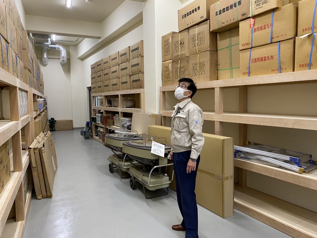 マスクを着用した作業着姿の市長が、天井まで届く棚にびっしりと段ボール箱が積まれた防災倉庫で、天井を見上げている写真