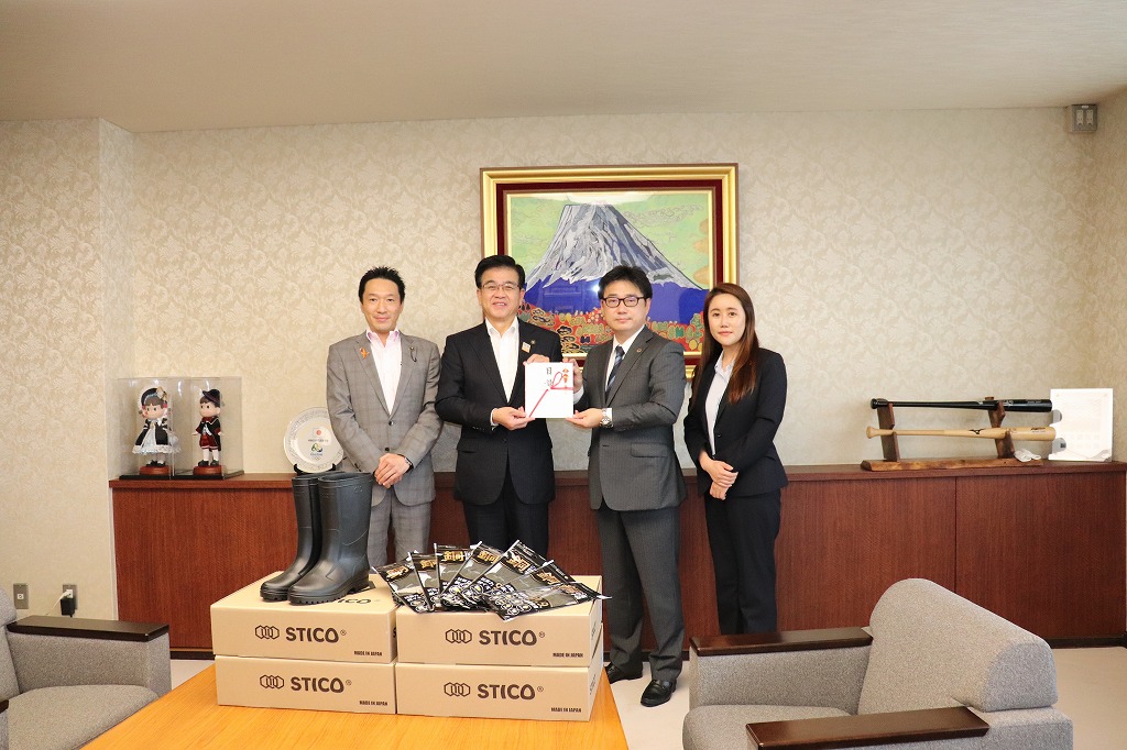 富士山の絵画が飾られた室内で、スーツ姿の市長と男性で目録を持ち、テーブルに積み上げられた長靴とSTICOと書かれた箱の寄贈品を囲んで男女4人が立っている写真