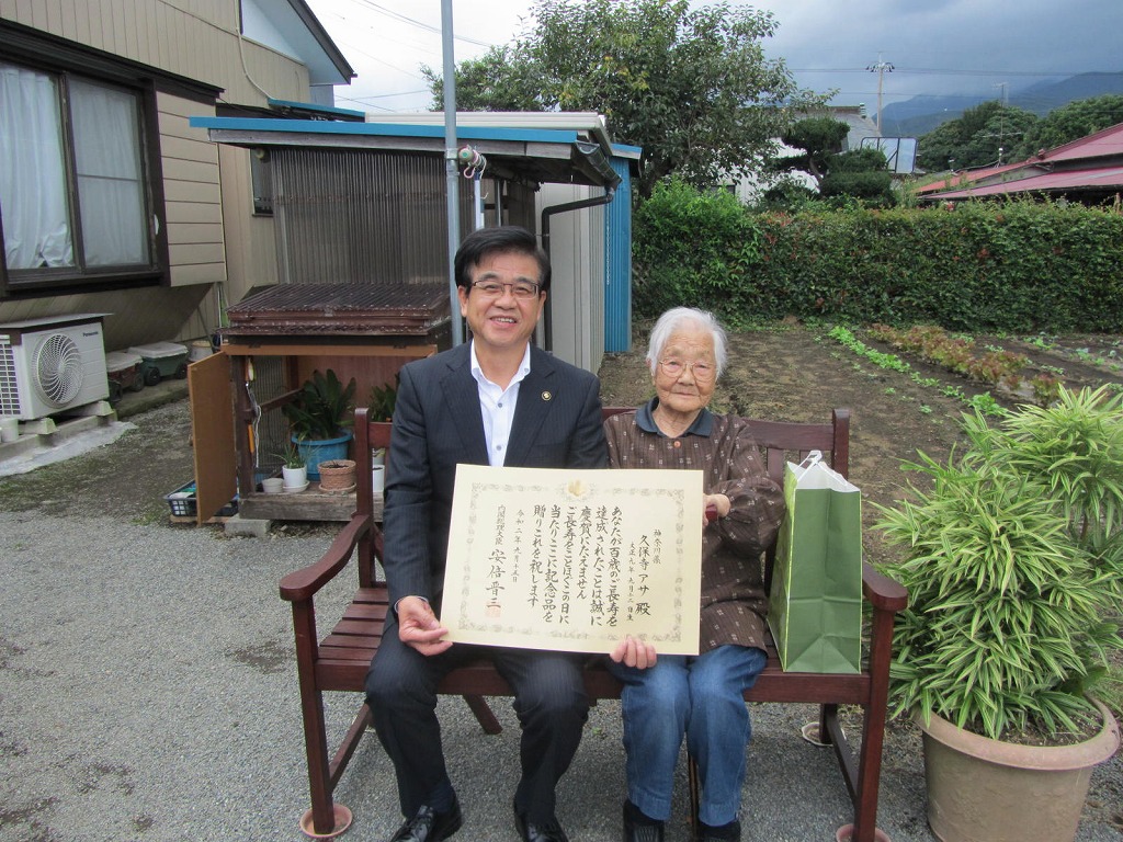 自宅の庭にある木製ベンチに座ったスーツ姿の市長と高齢女性が、長寿祝いの賞状と紙袋を囲んで笑顔で写っている写真