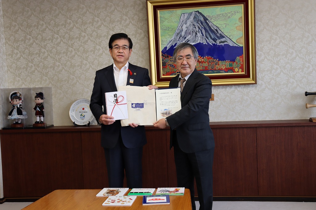 金色の額縁に入った山頂に雪が積もった大きな山の絵画が壁に飾られた落ち着いた背景の部屋で、コットンバックと目録を持ったスーツ姿の市長と書類を持った代表の男性が並んで立ち、カメラに向かい笑顔で記念撮影をしている写真