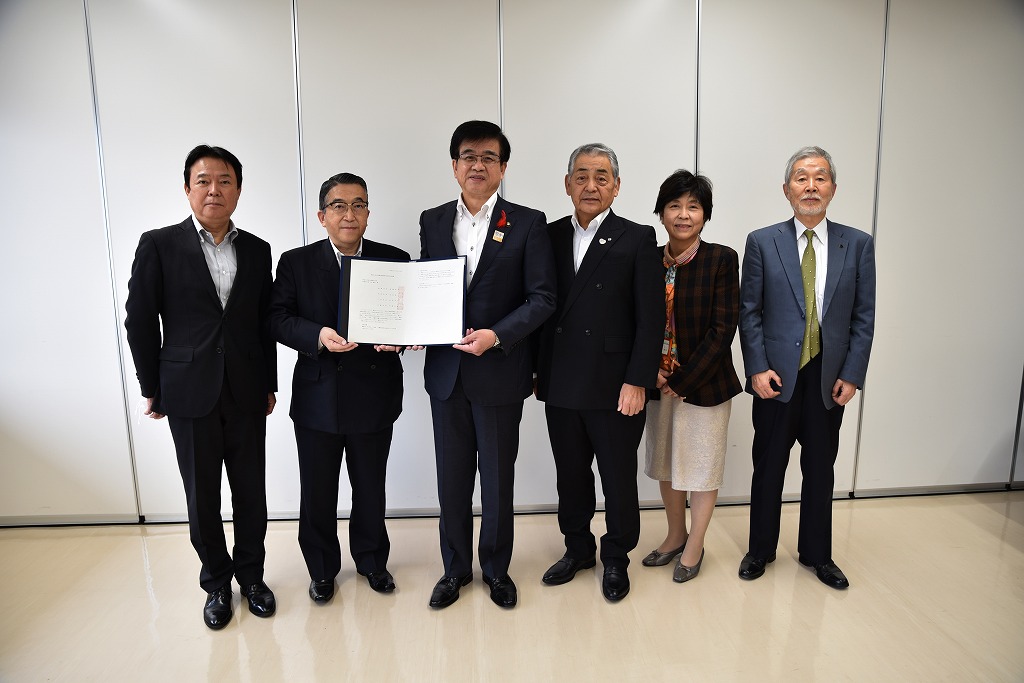 無地の白い壁の前でスーツ姿の男女6人が並んでおり、左から3番目に立つ市長と2番目に立つ代表の男性が書類を広げて記念撮影に応じている様子を捉えた写真