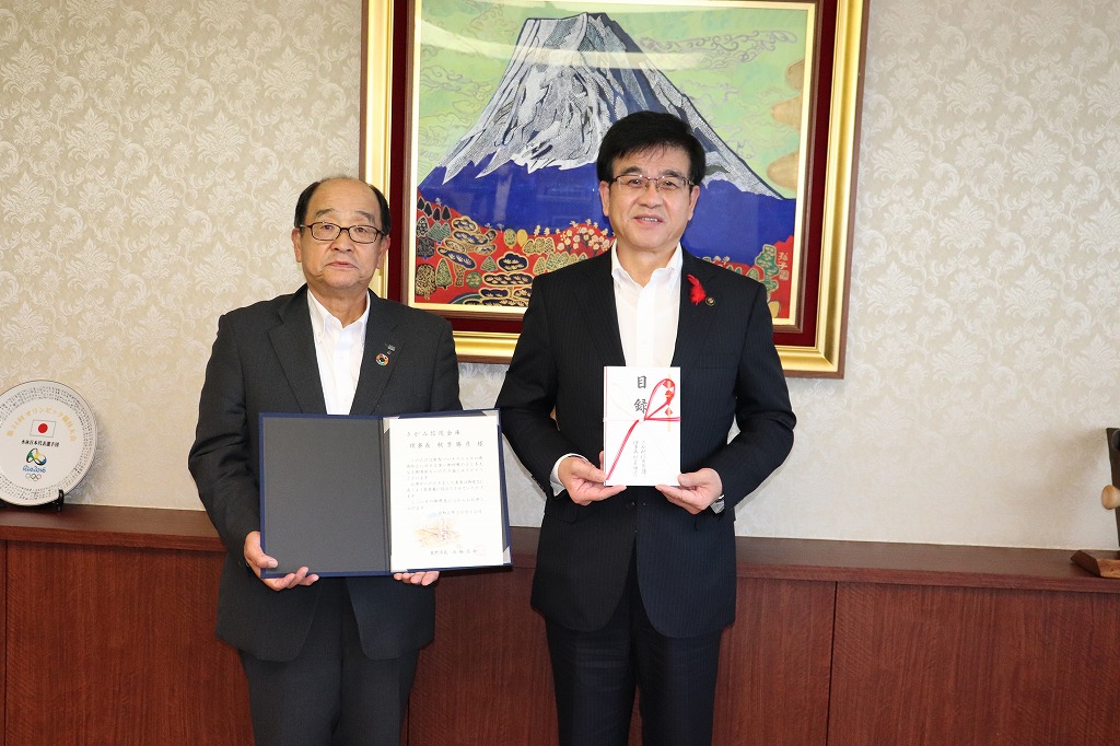 金色の額縁に入った山頂に雪が積もった大きな山の絵画を背景に、目録を持ったスーツ姿の市長と、書類を広げて持った秋葉理事長が並んで立ち、カメラに向かい記念撮影をしている写真