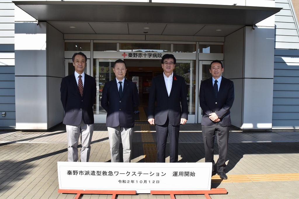 「秦野赤十字病院」と書かれた建物入り口の前にある「秦野市派遣型救急ワークステーション 運用開始令和2年10月12日」と書かれている白い看板が設置された前で、市長を含め4人のスーツ姿の男性が立ち並んでいる写真