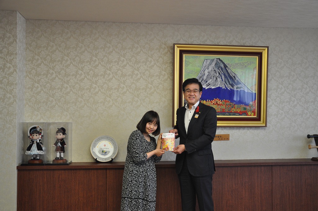 金色の額縁に入った山頂に雪が積もった大きな山の絵画が壁に飾られた落ち着いた市役所の応接室で、スーツ姿の市長と黒い花柄のワンピースを着た女性が、一冊の詩集を一緒に手に持って並び記念撮影に応じる様子を捉えた写真