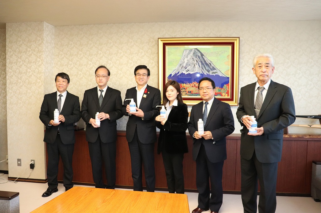 金色の額縁に入った山頂に雪が積もった大きな山の絵画が壁に飾られた落ち着いた背景の市役所の部屋で、市長を含めスーツ姿の男女6人が白いボトルを手に持って横一列に並び、記念撮影に応じている様子を捉えた写真