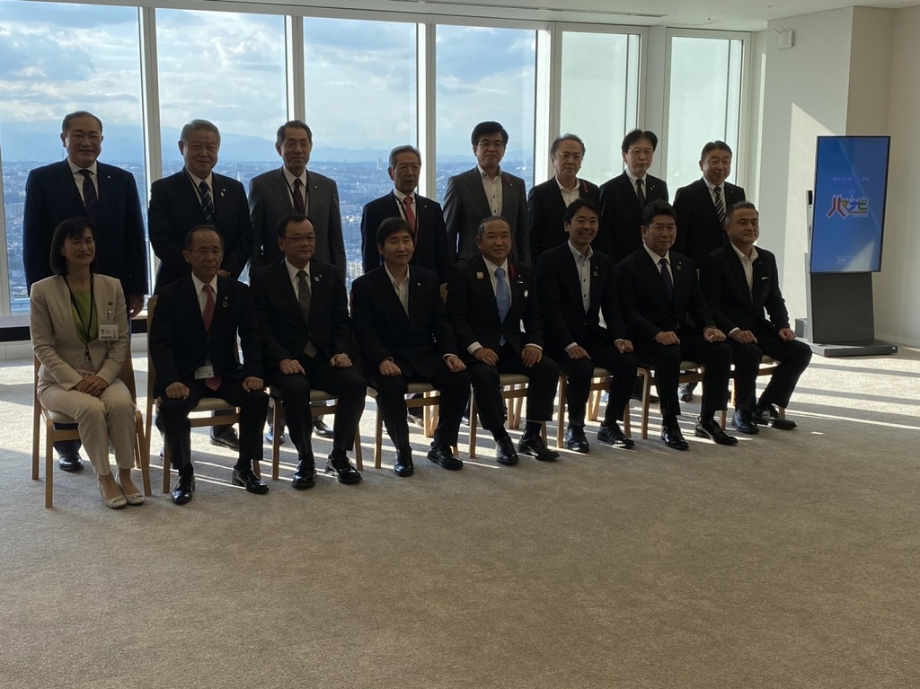 太陽の光が差し込む高層階の窓を背景に、スーツ姿の男女16人が前列の椅子に8人、後列に8人立ち、カメラに向かって並んでいる集合写真