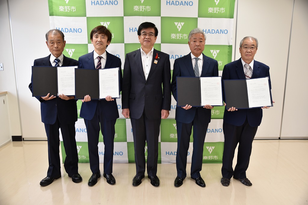 秦野市のバックパネルの背景を前に、スーツ姿の市長を中央に4人の男性が横一列に並び、それぞれが書類の入ったファイルを持って写真に収まっている様子を表した写真