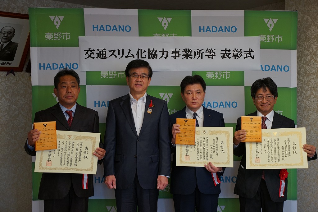 秦野市のバックパネルの前で、スーツ姿の市長と事業所代表の3人の男性が、一人ひとり表彰状とオレンジ色の記念品を手に持って記念撮影をしている写真