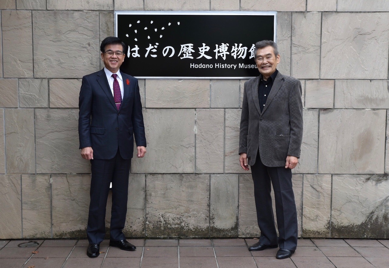 二人の男性が「はだの歴史博物館」と書かれた看板の前で笑顔で並んでおり、左の市長はスーツ、右の苅谷俊介さんはジャケットを着用している屋外での記念写真