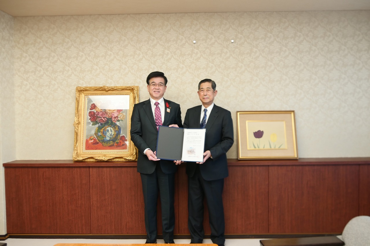 寄贈された二つの絵画の前で、スーツ姿の市長と男性が感謝状の入ったファイルを持ち、カメラに向かって微笑んでいる写真