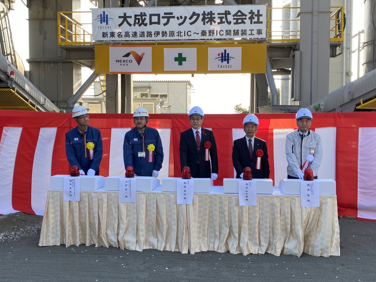 紅白の幕が張られた工場の屋外で、準備されたテーブルの前でヘルメットを被った5人の男性が並んでおり、上部には横断幕が飾られた安全祈念式典での火入れ式の写真