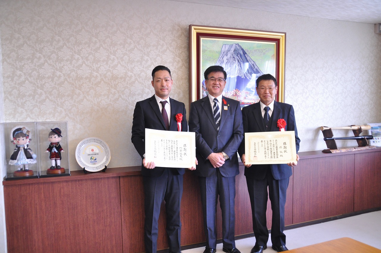 市の応接室でスーツ姿の市長を挟んでスーツ姿の男性2人が感謝状をもって並んでおり、カメラに向かって記念撮影をしている写真