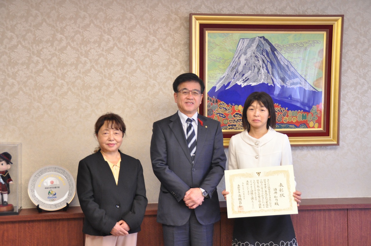 市の応接室でスーツ姿の市長を挟んで女性2人が並んでおり、向かって右側の女性が感謝状を持って微笑んでいる集合写真