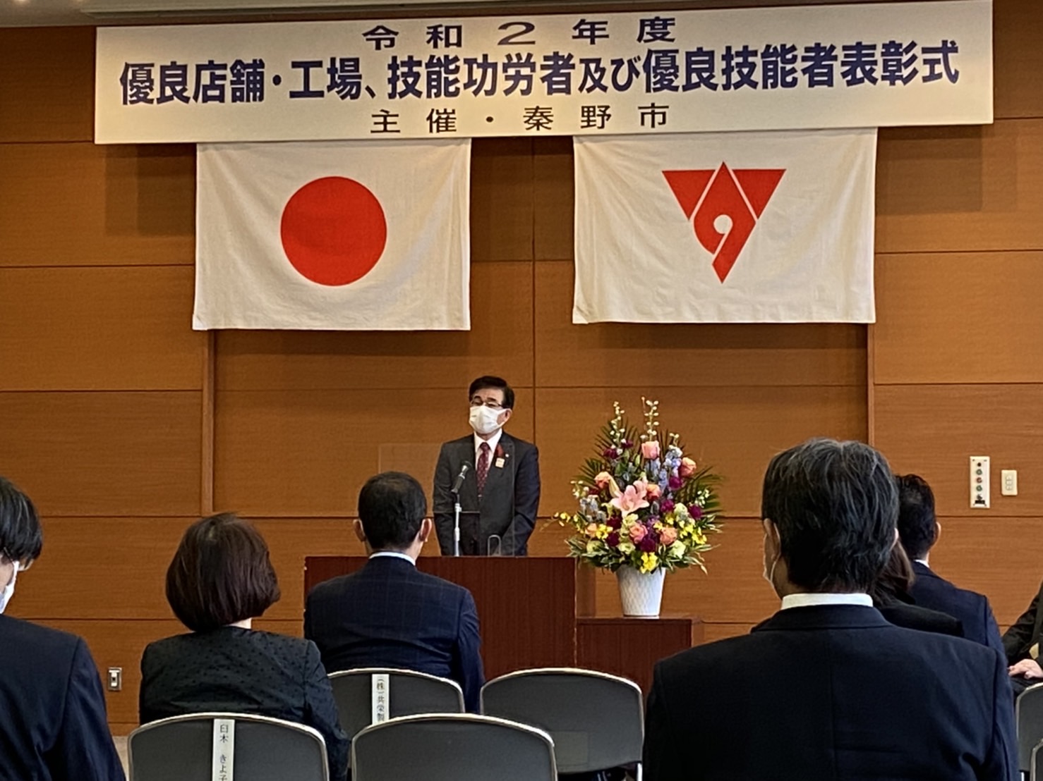 背景には日章旗と秦野市の市旗が掲げられている表彰式の会場でマスクをつけたスーツ姿の市長が、多くの聴衆を前に木製の演台から挨拶をしている写真