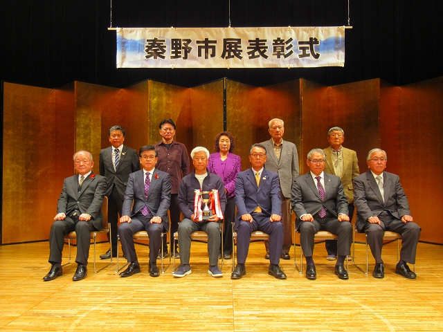 秦野市展表彰式と書かれた横断幕を背景に、スーツやジャケット姿の男性10名と女性1名がステージ上に並び、前列の椅子に座った6名の内左から3番目の男性がトロフィーを持ち、2列目の5名は立って記念撮影をしている写真
