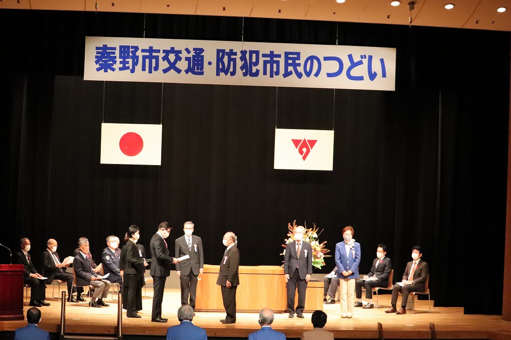 式典の壇上で複数の人が整列し、表彰が行われており、ステージ上部には「秦野市交通・防犯市民のつどい」の横断幕と日章旗が掲げられている写真