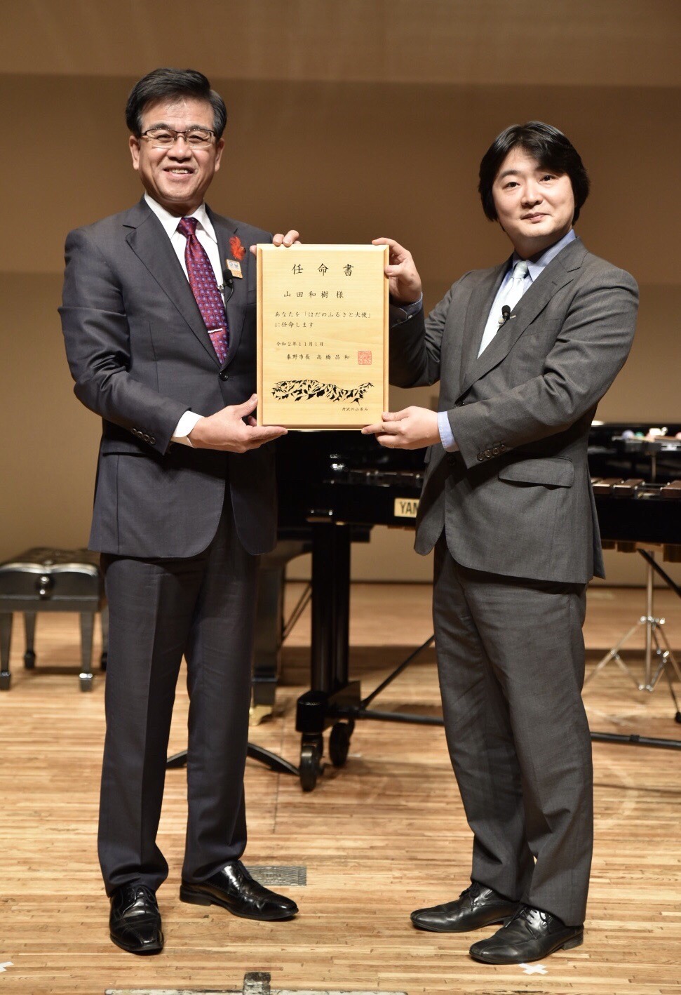 スーツ姿の市長と指揮者の山田和樹さんが任命書を両手でもってカメラに見せ記念撮影をしている写真