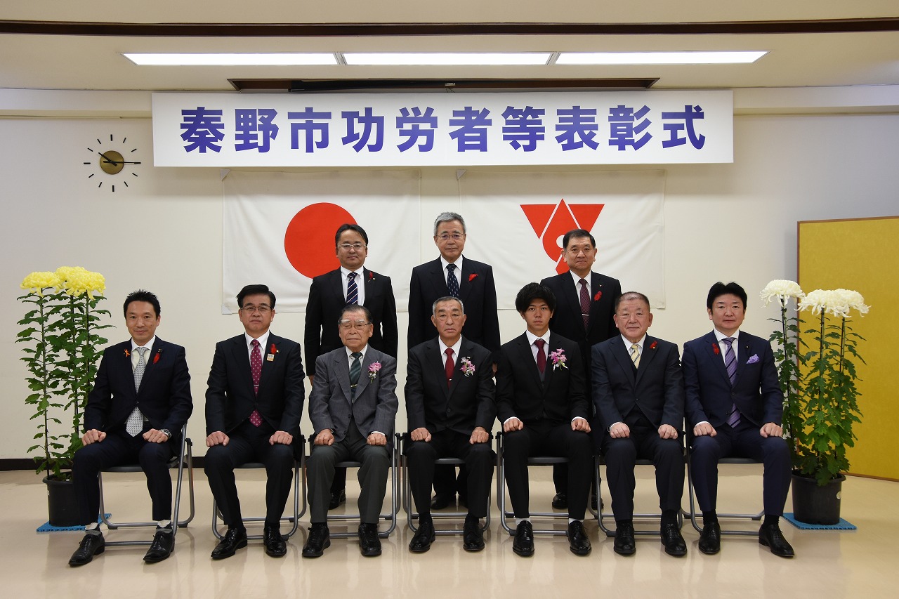 「秦野市功労者等表彰式」と書かれた横断幕を背景に、スーツ姿の男性たちが椅子に7名が座り、2列目に3名が立って並んでおり、両脇には鉢植えの黄色と白の花が飾られた表彰式の写真