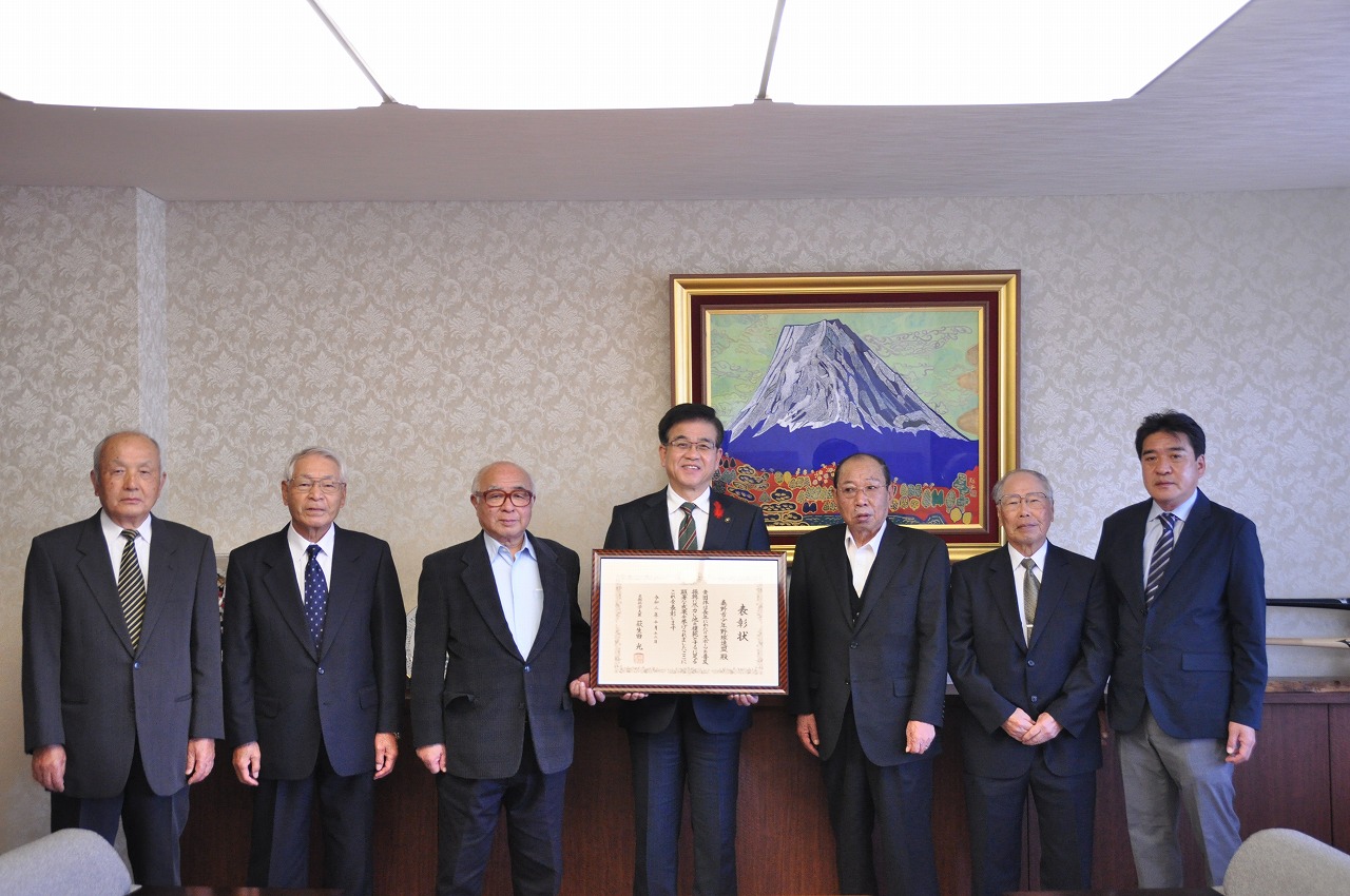 市役所の応接室で、スーツ姿の7人の男性がカメラに向かって並んでおり、中央の市長が感謝状の入った額縁を両手で持っている集合写真