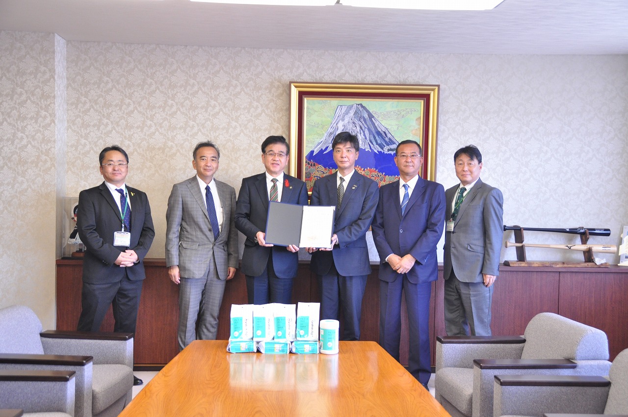 記念品の除菌クロスが置かれた机の後ろで、スーツ姿の男性6名が並んでおり、中央の市長と男性が感謝状をカメラに向かって見せている室内での写真