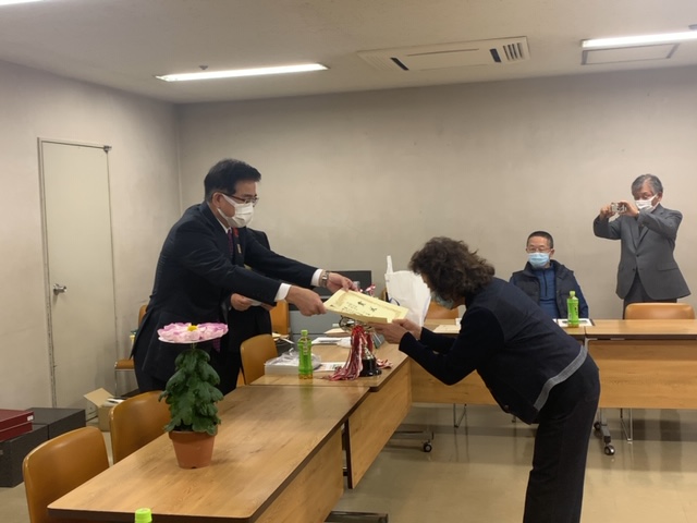 感謝状のようなものを手渡す市長と、それを受け取る女性が部屋の中央に立っており、奥のテーブル席では別の男性がスマートフォンでその様子を撮影している授賞式の写真