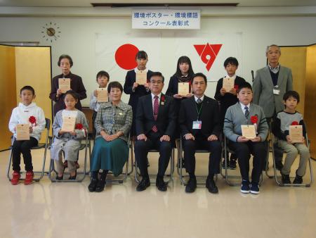 「環境ポスター・環境標語コンクール表彰式」と書かれた横断幕の下で、スーツ姿の男性を中央に子供から大人までが賞状を持って並び、記念撮影をしている写真
