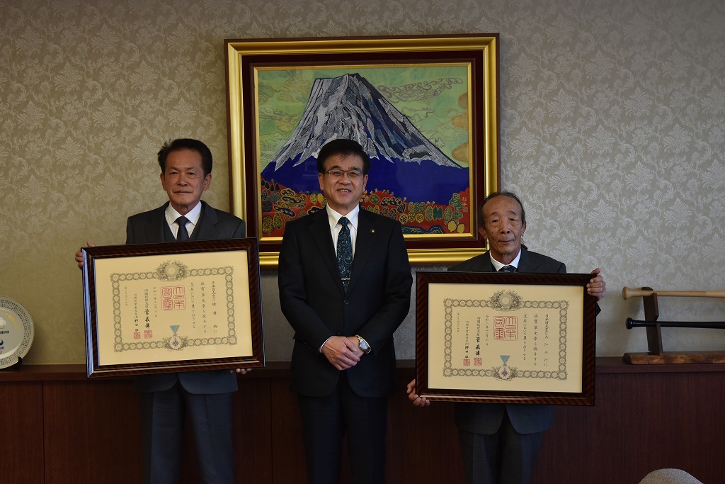 富士山の絵画が飾られた部屋で、中央の市長は笑顔で、両脇の男性は額縁入りの表彰状を持って立っている写真