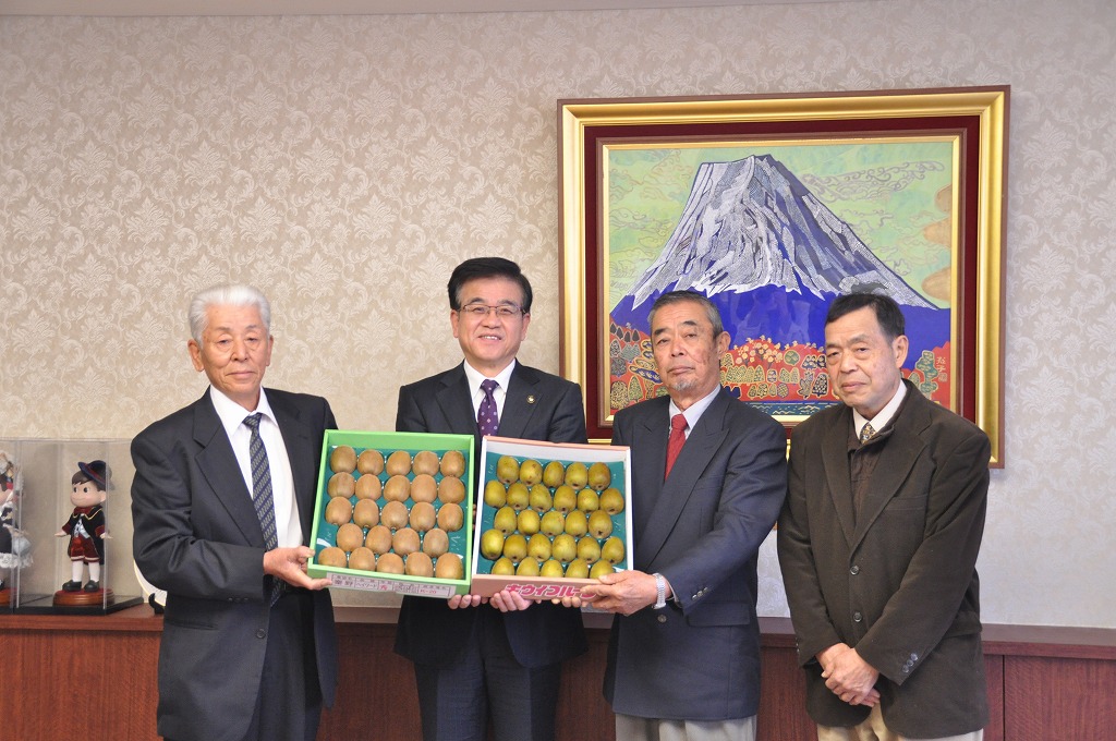 富士山の絵画が飾られた室内で、市長を含めたスーツ姿の4人の男性が、それぞれ緑と黄色のキウイフルーツの箱を持って立っている写真