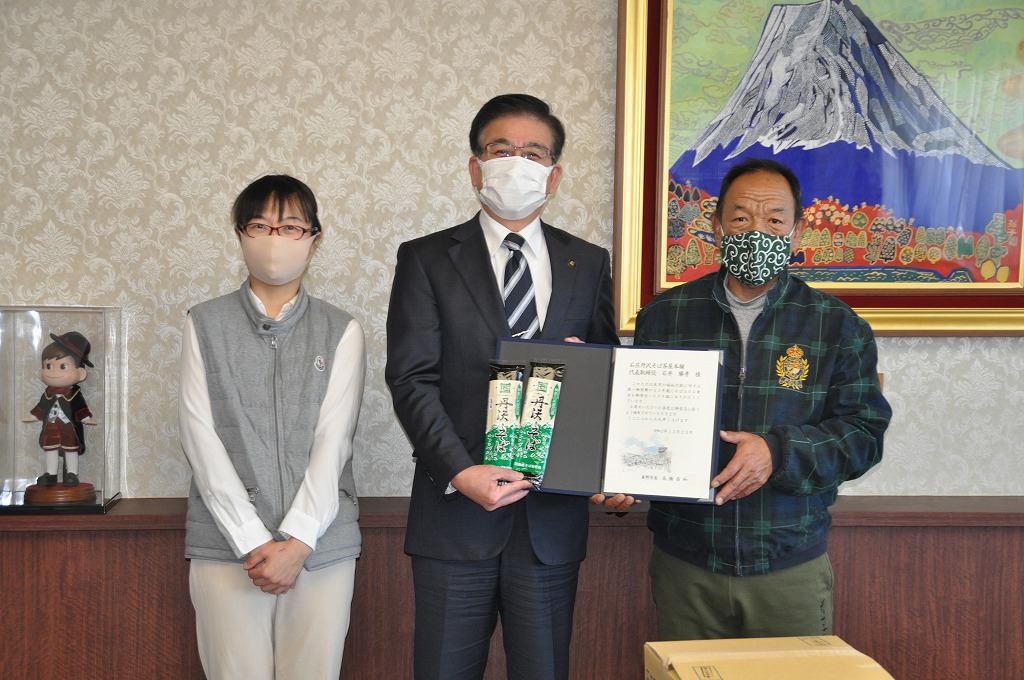 富士山の絵画が飾られた部屋で、真ん中の市長は袋詰めされた蕎麦、右側の男性は感謝状のような書類を持ち、マスクを着用した3人の男女が並んで写っている写真