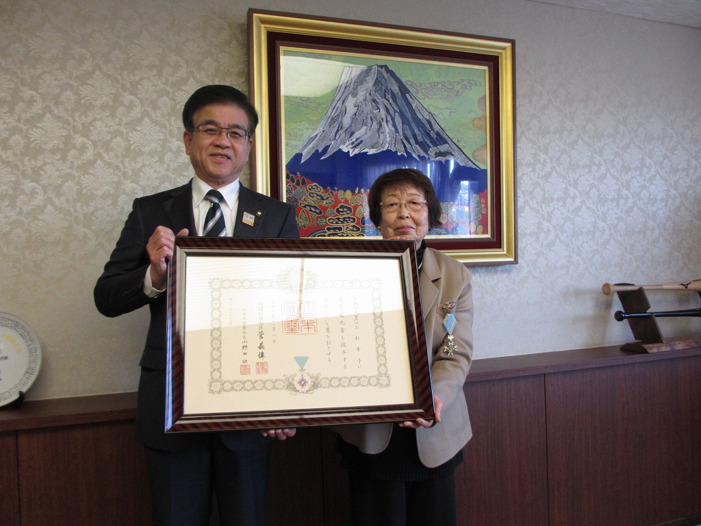 富士山の絵画が飾られた部屋で、スーツ姿の市長と、勲章をつけた高齢の女性が、大きな額縁に入った感謝状を両手で持って並んでいる写真