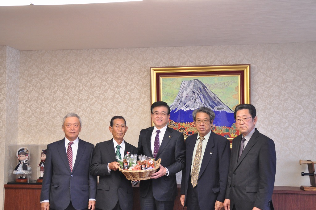富士山の絵画が飾られた室内で、スーツ姿の5人の男性が並んでおり、中央の市長は籠いっぱいの寄贈品を持って笑顔で立っている写真