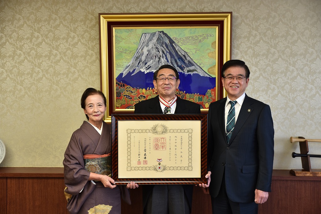 富士山の絵画が飾られた部屋で、和服姿の女性とモーニング姿の男性とスーツ姿の市長が、中央で勲章のついた大きな表彰状を挟んで、笑顔で記念撮影している写真