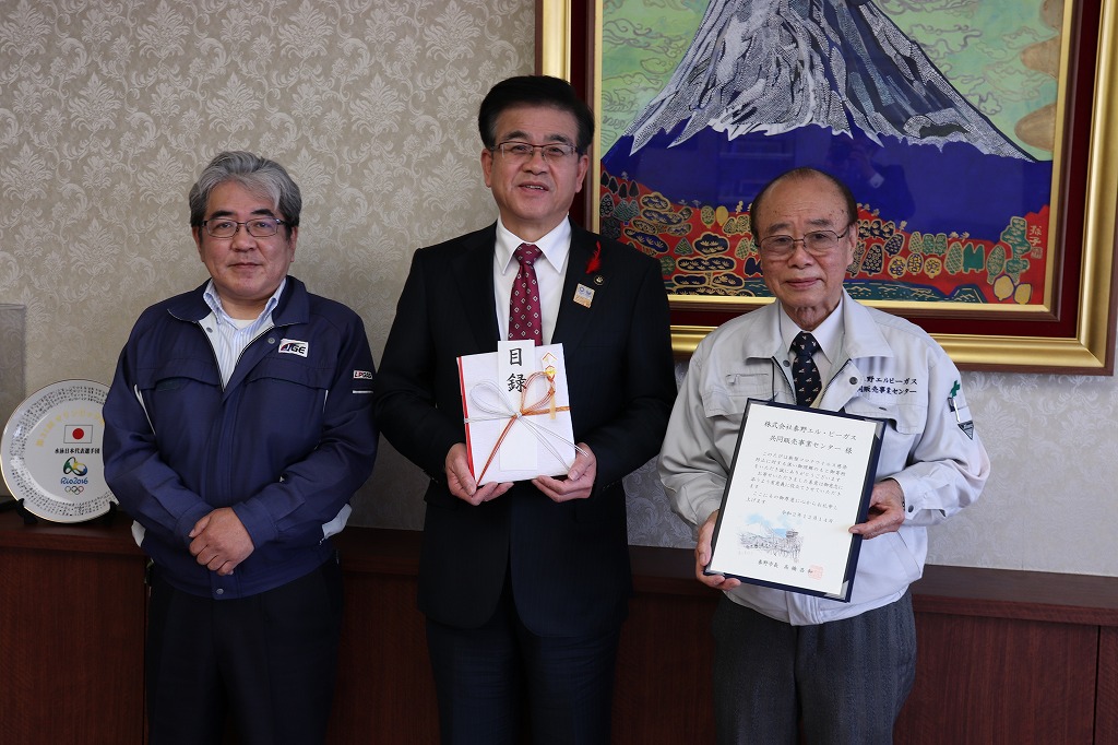 富士山の絵画が飾られた部屋で、スーツ姿の市長が目録を持ち、作業服姿の二人の男性が感謝状のような書類を持って並んでいる写真