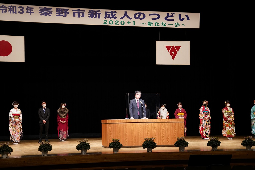「令和3年秦野市新成人のつどい」と書かれた横断幕のステージ上で新成人たちが和装やスーツを着用して並んでおり、市長がアクリル板越しの演壇からスピーチをしている様子の写真