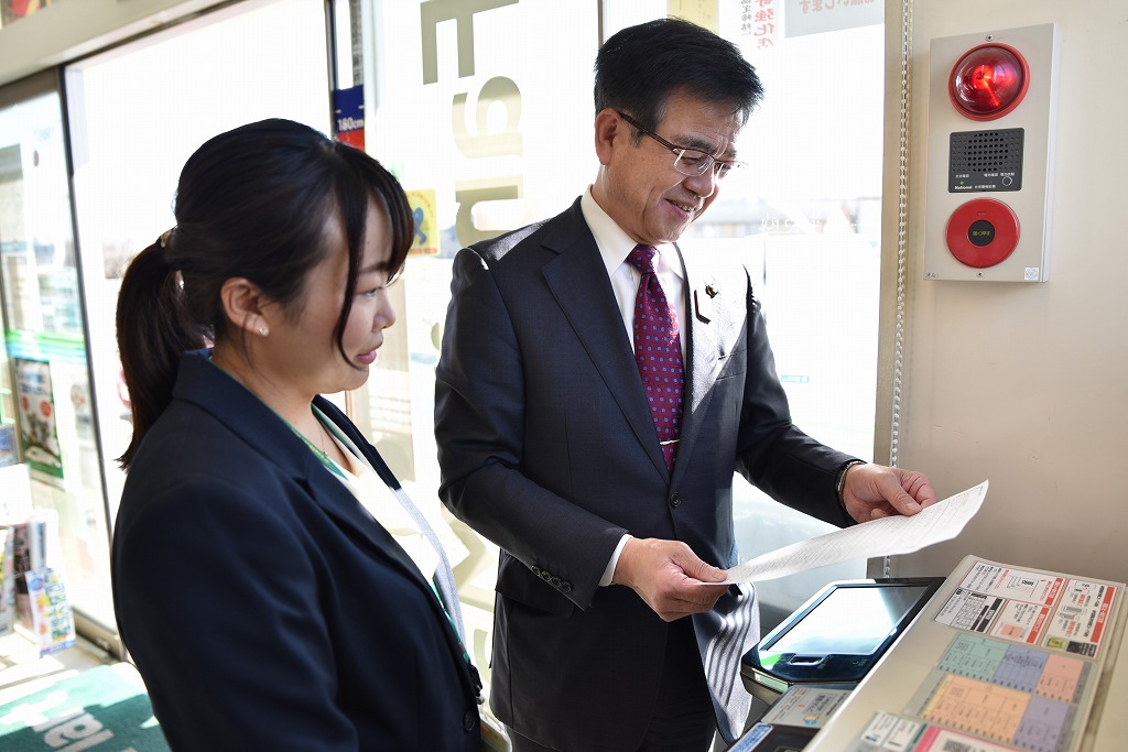 コンビニのコピー機の前で市長と女性が並んで立ち、交付サービスを試している様子の写真