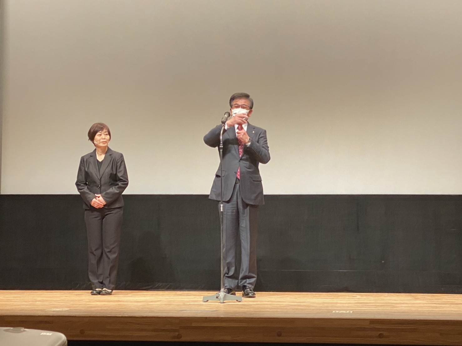 ステージ上で、スーツ姿の市長がマイクに向かって手話を交えてスピーチをしており、その横にスーツを着た女性が立って見守っている写真