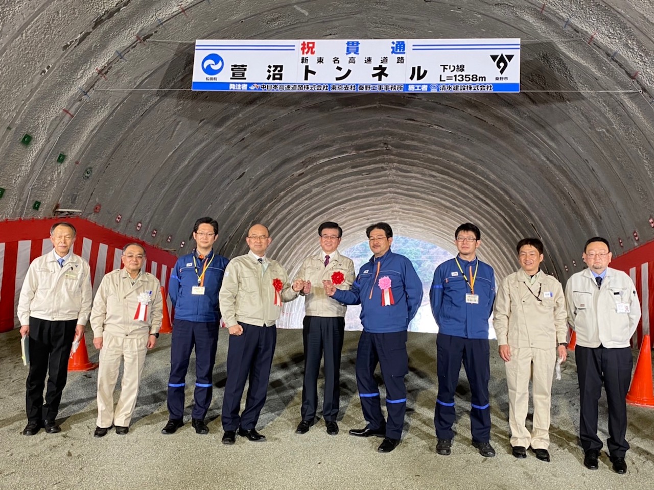 新東名高速道路のトンネル内で、複数の作業服姿の男性が並んでおり、中央には「祝 貫通 葛葉トンネル」と書かれた看板が掲げられている記念写真
