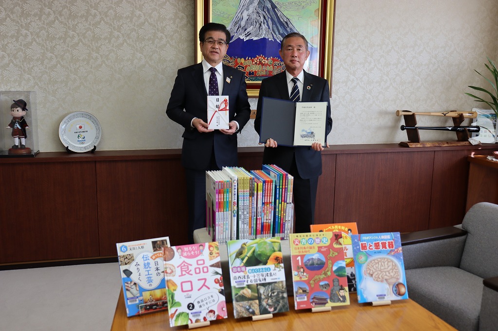 室内の机の上に複数の子ども向け図書が立てて並べられており、その後ろに立つ市長は金封を持ち、男性が賞状を持っている写真