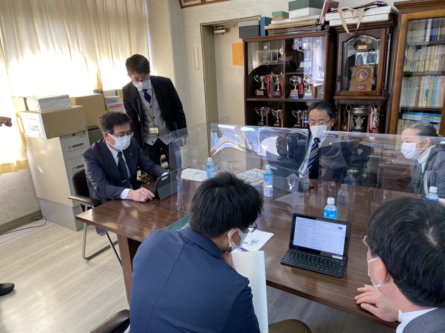 市長と関係者の人々がテーブルを囲んで座っており、アクリル板で仕切られた中でタブレットを操作しながら話し合っている様子の写真