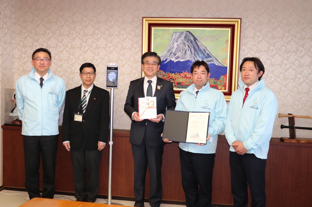 富士山の絵画が飾られた部屋で、水色のブルゾンを着た3人とスーツ姿の男性と市長が横並びで金封や感謝状を持ち、市長の横にサーマルカメラが設置し記念撮影している写真写真